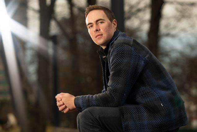 Photo: Cameron Monesmith, a white man with short hair wearing a flannel shirt, sits outside, his left side profile to the camera, hands clasp, posing for the photo.