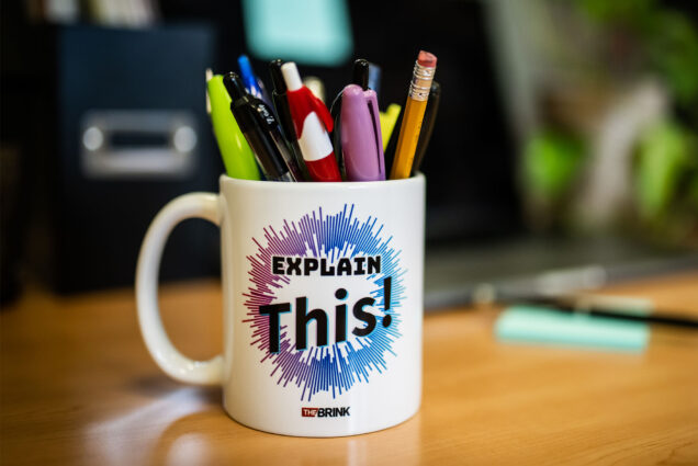 Photo: A white mug with a logo overlay reading EXPLAIN THIS! The text is featured over a burst design of purple and blue. Various pens and pencils are in the mug. The blurred background depicts office-type material.