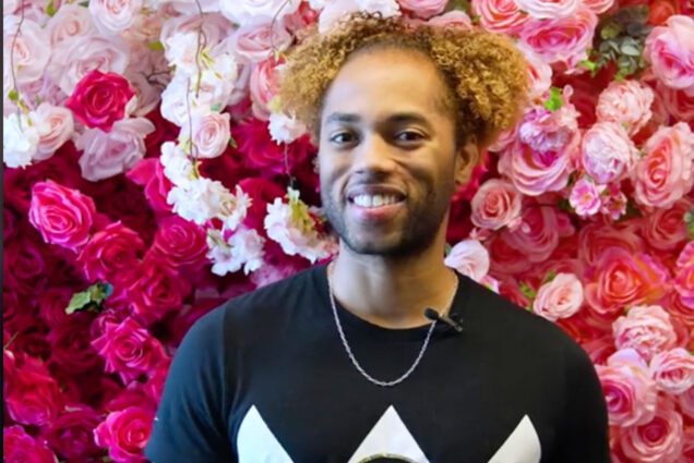 Photo: A young black man standing in front of a background of roses with a smile on his face
