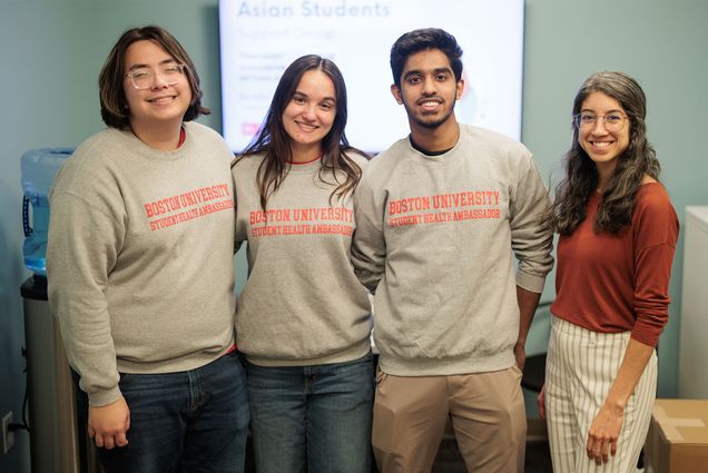 Photo: A group of students wearing matching Boston University sweatshirts smile for a picture alongside their faculty partner, a woman with long curly hair, glasses and a red shirt