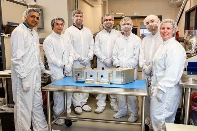 Photo: A group of scientists in white outfits and hair nets stand around a large metal machine with proud looks on their faces in a lab setting