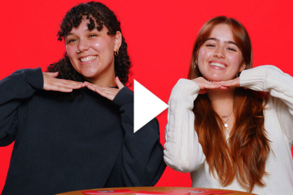 Photo: Two college students in front of a red background smile and hold their hands under their chins. There is a white play button overlayed on top of the image
