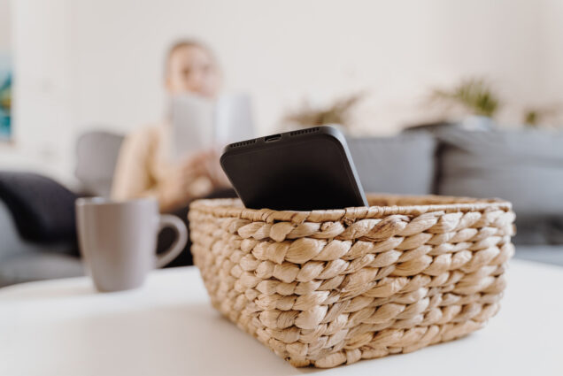 Photo: A woman sits on a couch reading a book while her phone is in a basket on a nearby table