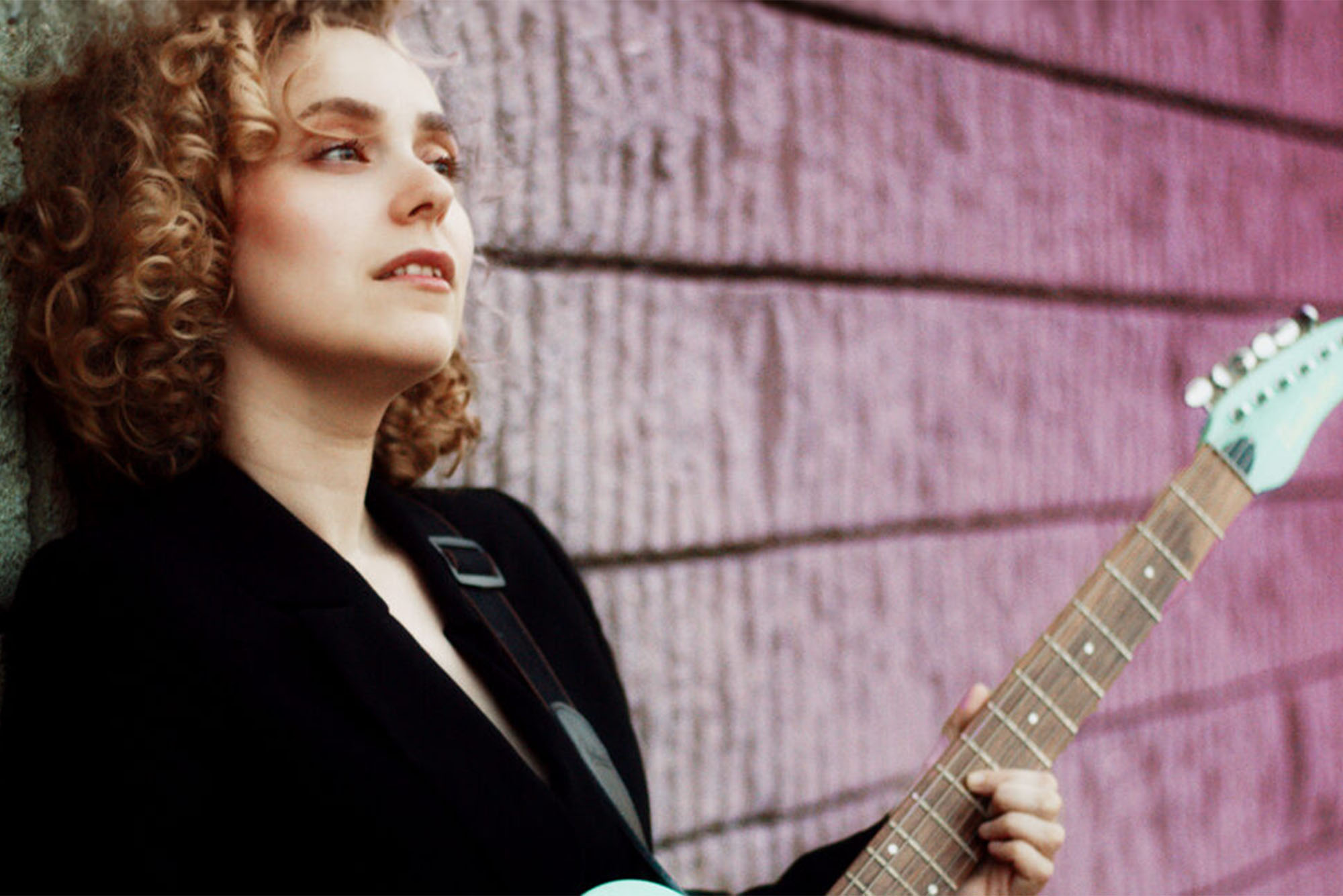 Photo: A person holding an electric guitar with curly hair in front of a brick wall