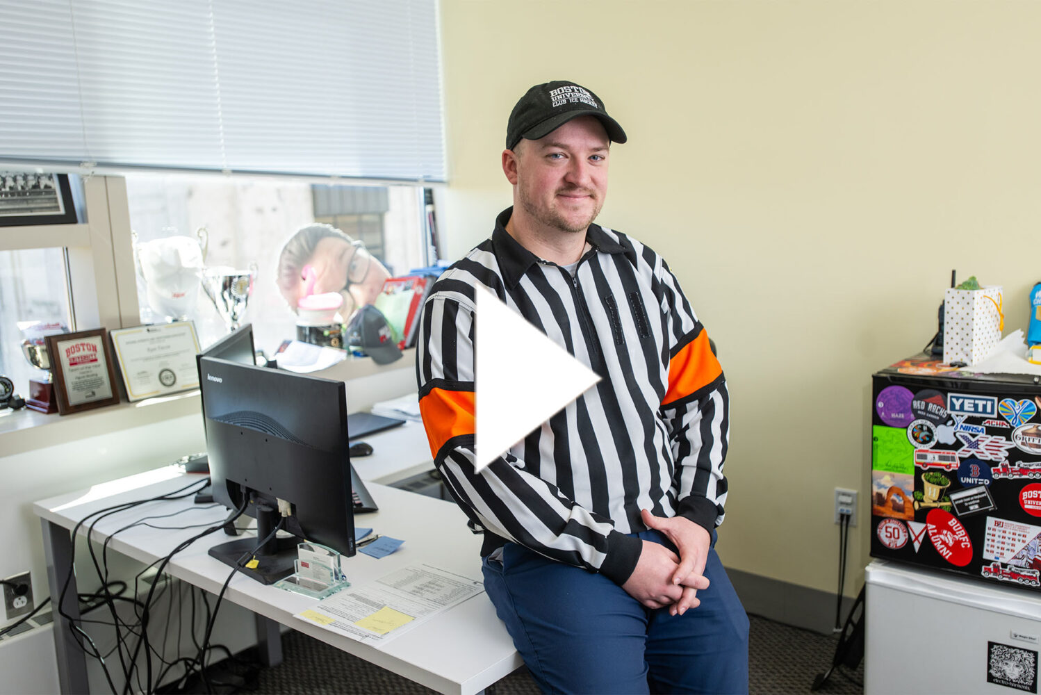 Photo: A man in a referee outfit with a hat and a long-sleeved shirt with vertical black and white stripes. There is a white play button in the center of the image