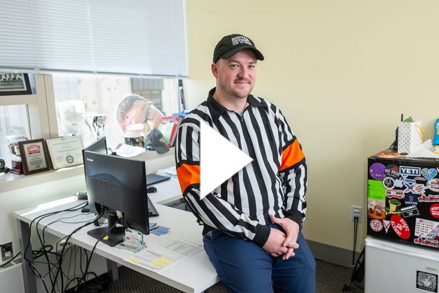 Photo: A man in a referee outfit with a hat and a long-sleeved shirt with vertical black and white stripes. There is a white play button in the center of the image