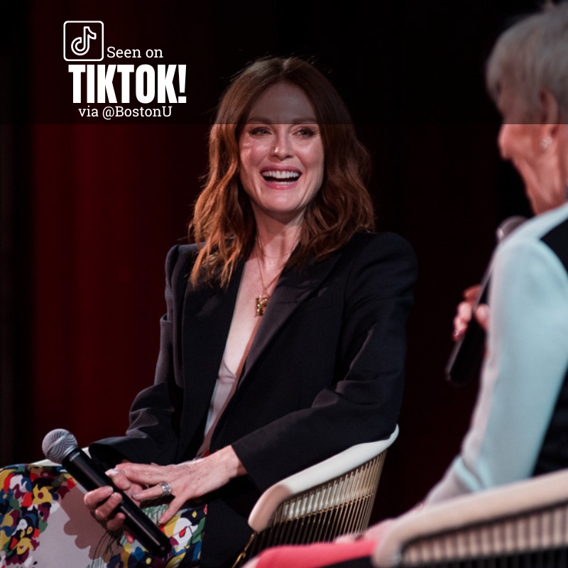 Photo: Julianne Moore, a white actress smiles in response to a panel talker. Moore faces the woman to her right, said woman has short hair and a blue top. Moore wears a black blazer and floral pants.