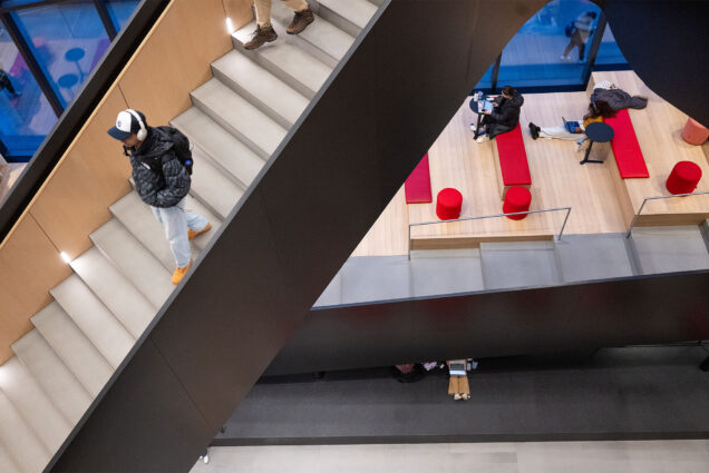 Photo: A college student walks down a staircase as other students study on the ground floor below.