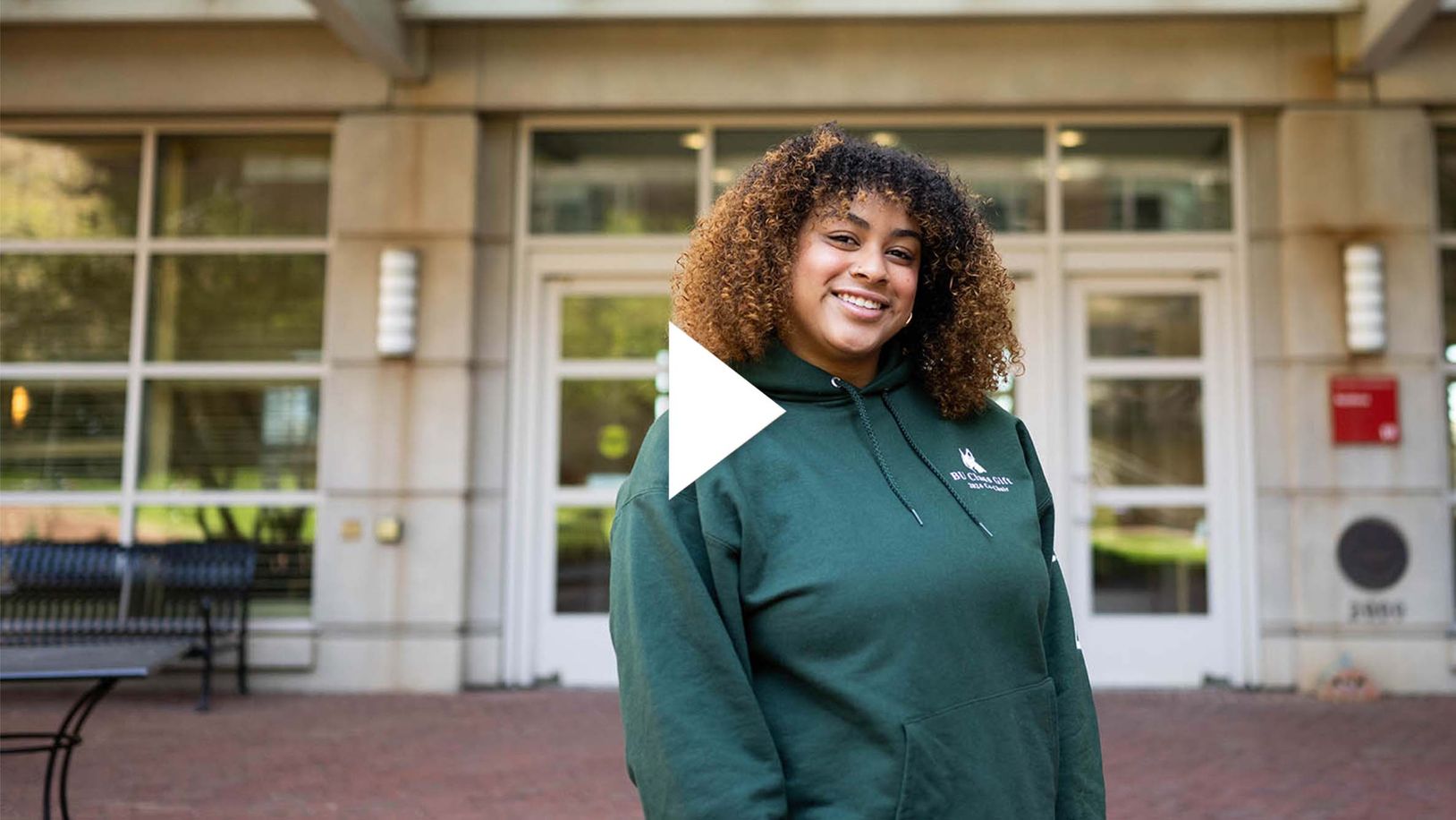 Photo: Yadira Cabrera, a young woman with shoulder length curly hair and bangs, at StuVi dorm buildings front door. A play button overlay rest in the middle of the photo.