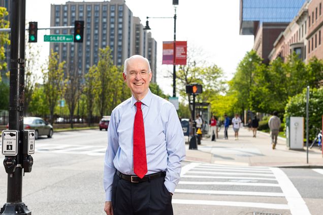 Photo: Kenneth Freeman, a white man wearing glasses, a light blue collared shirt, red tie, and black pants smiles and poses on a sidewalk on Commonwealth Avenue. Trees and various BU buildings can be seen in the blurry background on the sunny day.