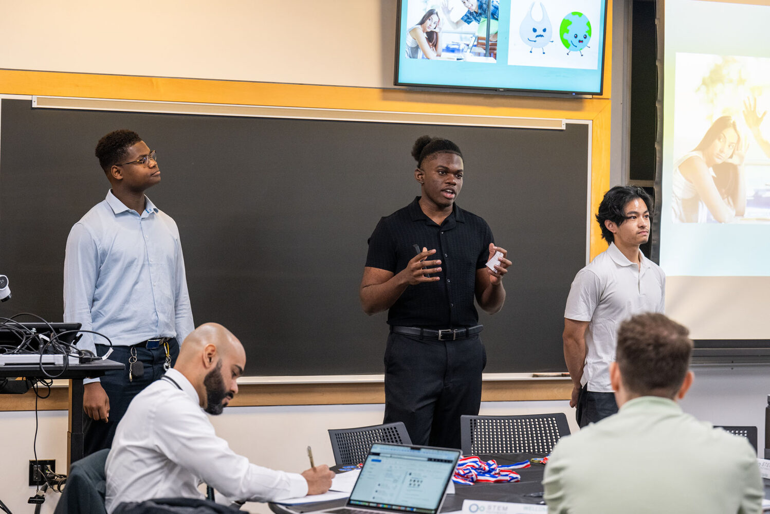 Photo: Three young men stand at the front of a classroom presenting their pitches. On the left, a Black man with a flat top fade wears a blue button up and navy slacks. In the middle, a Black man with his hair pulled into a puff wears a black button up with black slacks. And on the right, a young Asian man with long hair wears a gray polo and slacks.