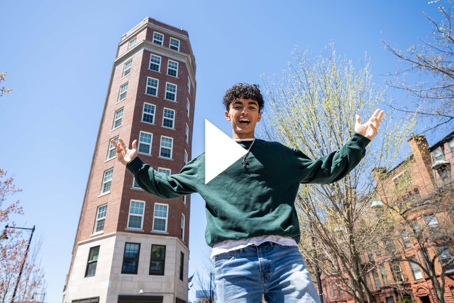 Photo: A picture of a male student posing in front of a dorm building in the city with his arms extended. There is a video play button over the image