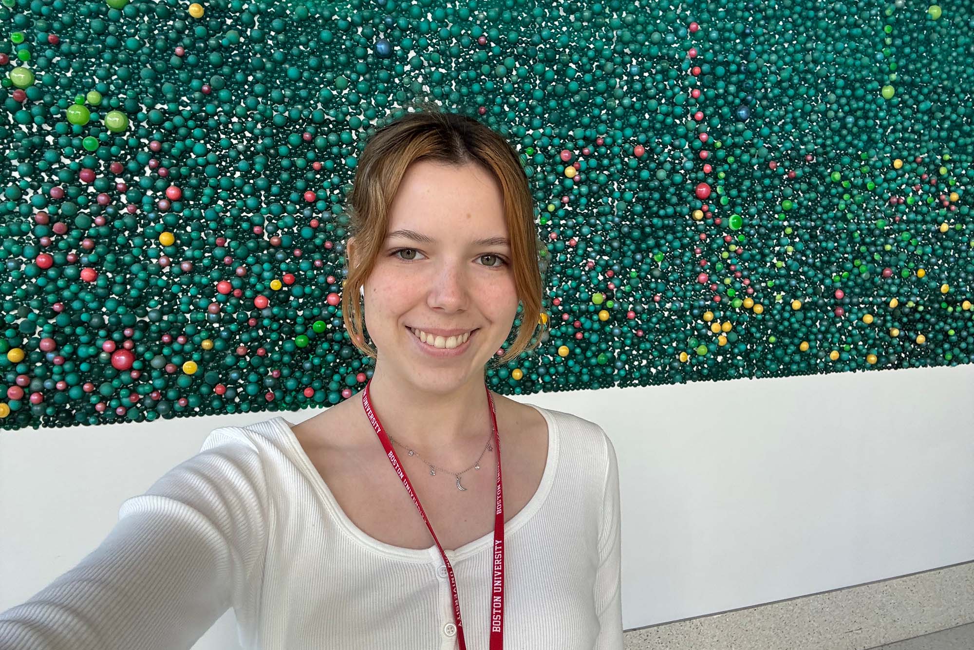 Photo: A woman with blonde hair smiling in front of a public art mural while wearing a long-sleeved white shirt and a Boston University lanyard