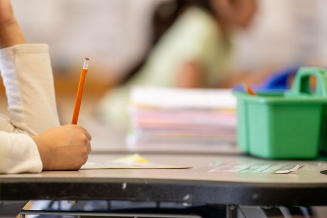 Photo: A close-up of an elementary student writing with a pencil with school supplies on their desk