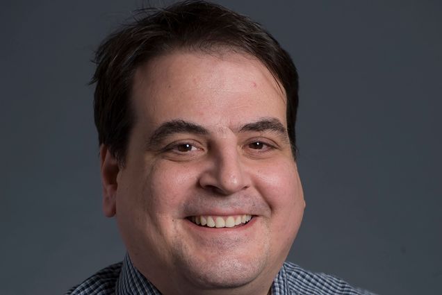 Photo: A headshot of Joseph Massaro, a white man smiling into the camera. He wears a blue and grey, tight checkered button up shirt.