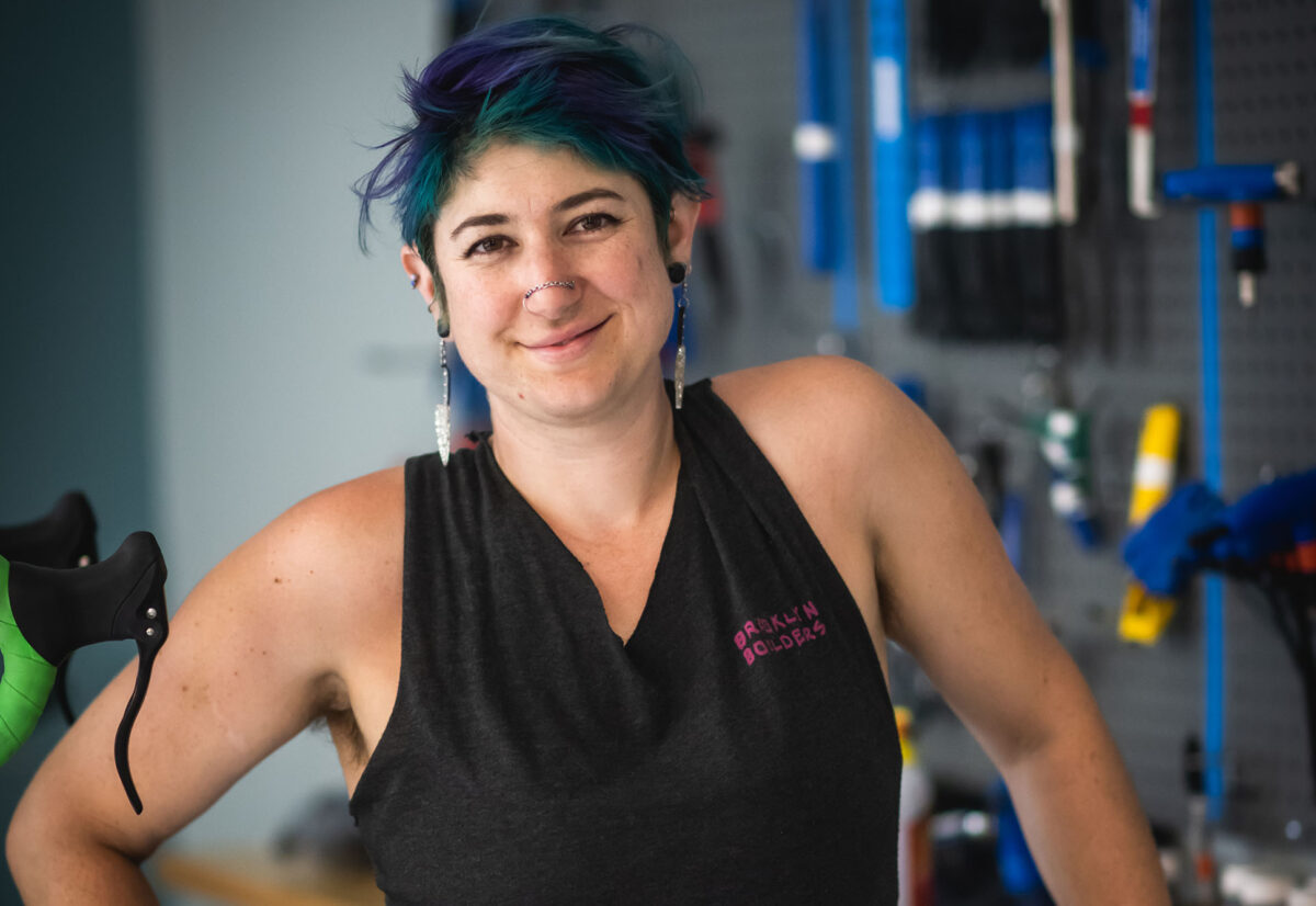 Jay Diengott, manager of BU's new Bike Kitchen, smiles for the camera. They have blue, green hair, dangling earrings, and a double nose piercing.