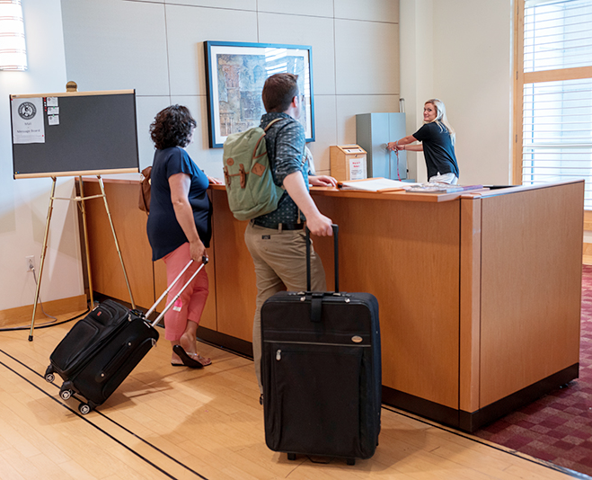 Two people with suitcases checking in at front desk with student worker behind it.