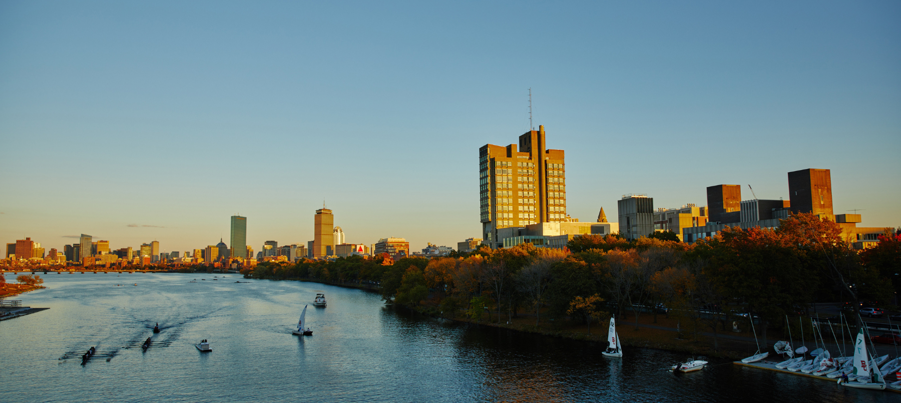 A view of the Charles River and Boston University campus in golden, late afternoon sunlight. There are several boats on the river and you can see many BU campus buildings and the Boston skyline is in the background