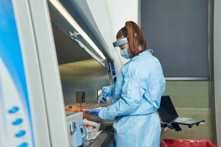 A person wearing a face shield, wearing a lab gown and gloves works in a hood.
