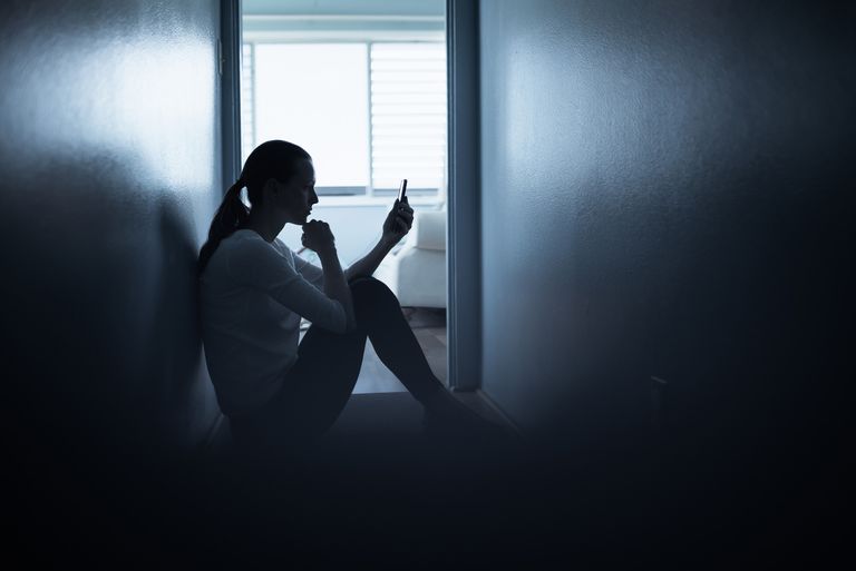 A woman sits on the floor staring at her phone in a dark narrow hallway