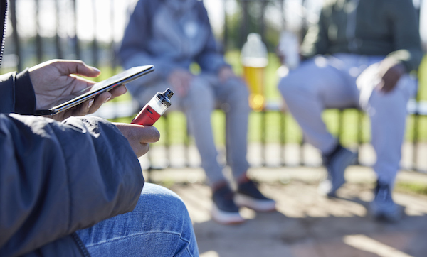 Close Up Of Teenagers With Mobile Phone Vaping and Drinking Alcohol In Park