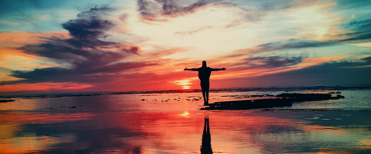 Person standing on the beach at sunset, arms stretched wide.