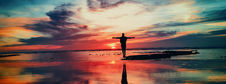 Person standing on the beach at sunset, arms stretched wide.
