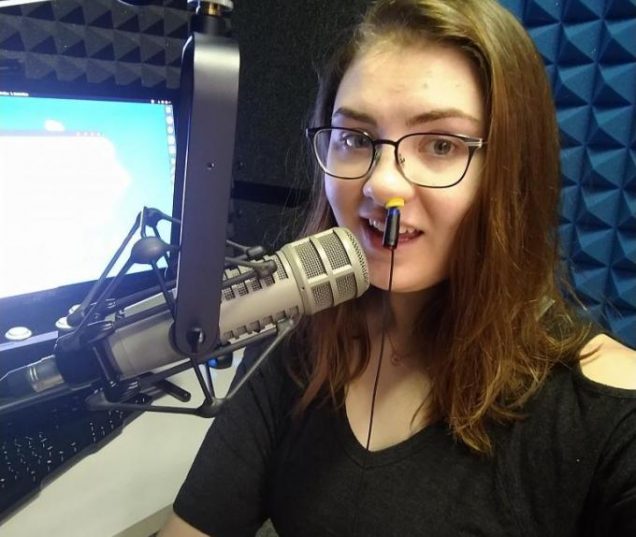 Aleah Combs, a white female with long hair and glasses, speaks into a microphone in a sound booth with a velopharyngeal tracer in their nose.