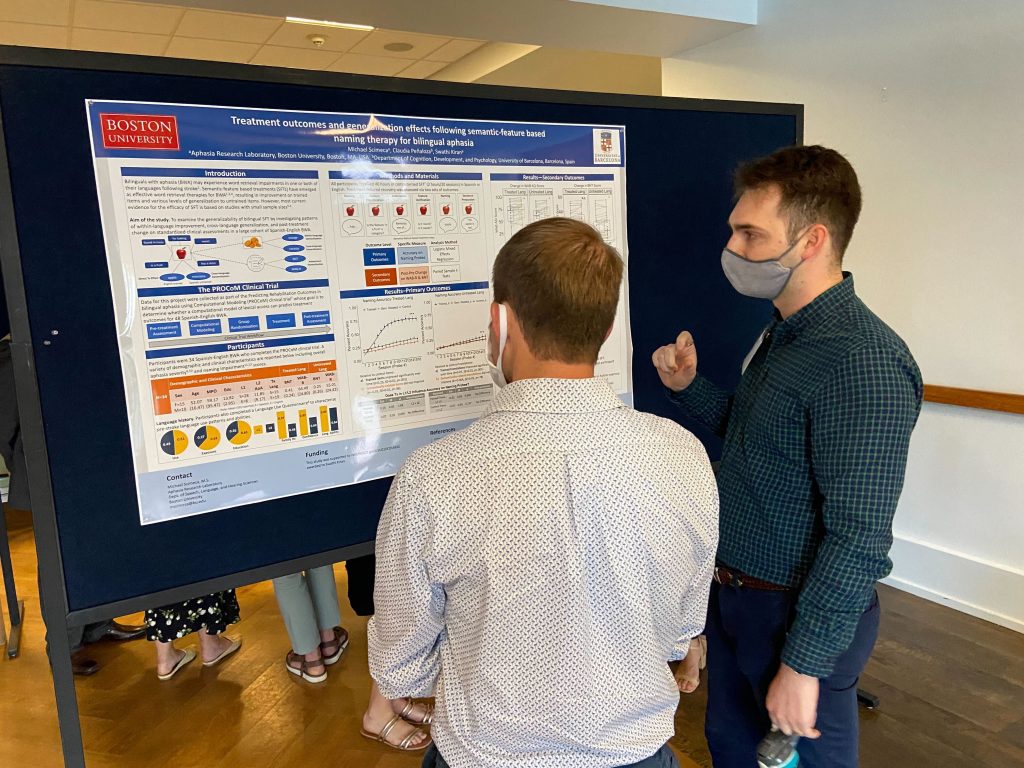 Two people with short hair, masks, and button-down shirts stand in front of a poster that is titled "Treatment outcomes and generation effects following semantic-feature based naming therapy for bilingual aphasia".