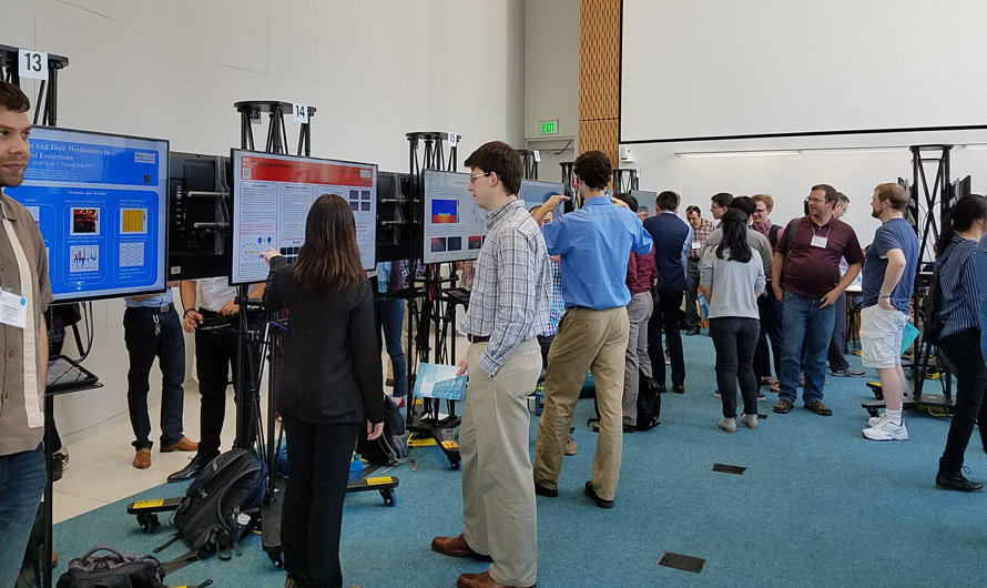People attending a symposium gathered around different electronic screens