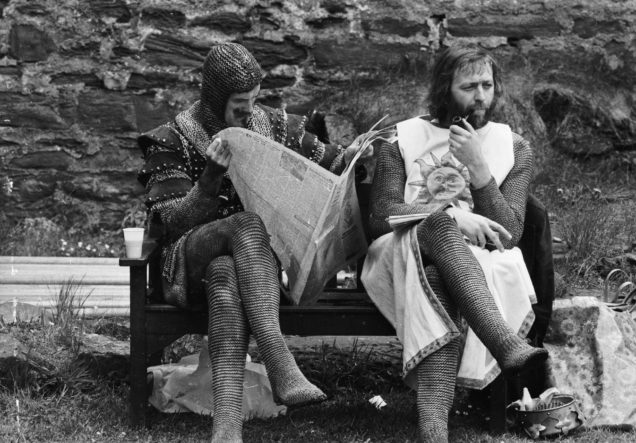 21st May 1974: A chainmail-clad John Cleese reads a newspaper while Graham Chapman smokes a quiet pipe on the set of 'Monty Python and the Holy Grail'. (Photo by John Downing/Express/Getty Images)