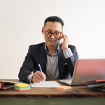 A man sits at his desk while on the phone, looking at his laptop, and read to write with a piece of paper and pen in hand.