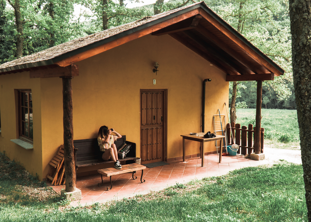 A girl is sitting on the front porch of a yellow house, which is located in the woods, and is taking a photograph.