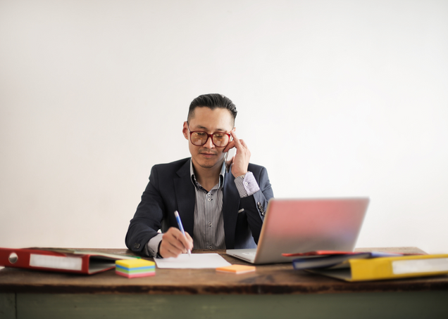 A man sits at his desk while on the phone, looking at his laptop, and read to write with a piece of paper and pen in hand.