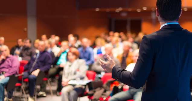 In focus we see the back of a man speaking at a conference to a crowd, which is blurred in the background