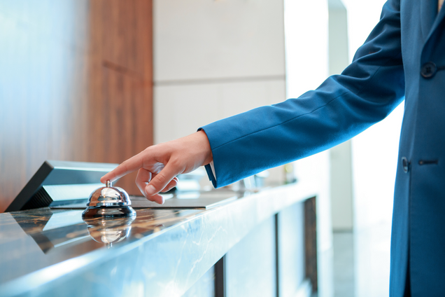 arm of person wearing a blue blazer reaches to press a bell on top of hotel lobby counter