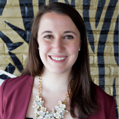 Profile image of Maura Feltault in maroon blazer and wearing a floral necklace against a painted brick background