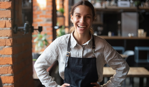 Cheerful young waitress wearing apron laughing looking at camera, happy businesswoman small business owner of girl entrepreneur cafe employee posing in restaurant coffee shop interior, portrait