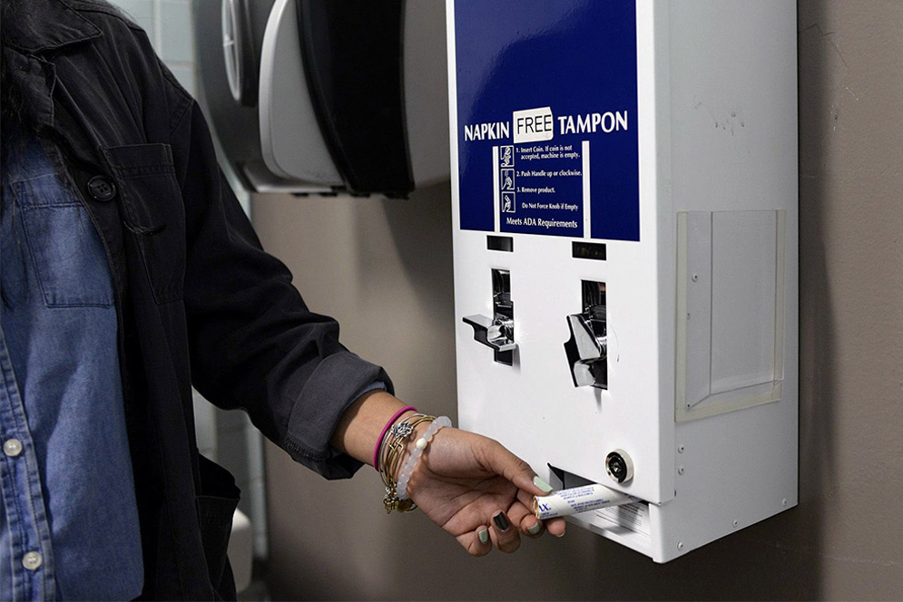 A young woman removes a tampon from a blue and white free tampons dispenser in a restroom.