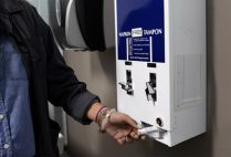 A young woman removes a tampon from a blue and white free tampons dispenser in a restroom.