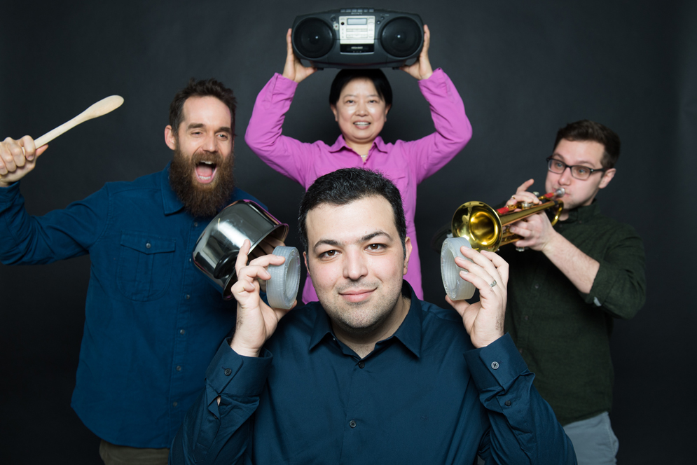 Boston University Ph. D. candidate Reza Ghaffarivardavagh poses with his research team. Reza holds the noise cancellation devices his team developed over his ears while his teammates make noise banging a pot, playing a trumpet, and a boombox in the background