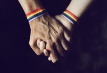 closeup of two caucasian men holding hands with a rainbow-patterned wristban on their wrists