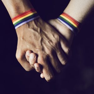 closeup of two caucasian men holding hands with a rainbow-patterned wristban on their wrists