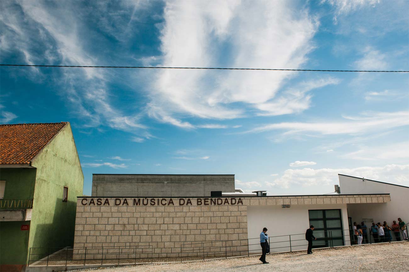 Full color slideshow photo of an exterior view of the entrance to the Casa Da Musica Da Bendada music school with Bendada Music Festival attendees outside.