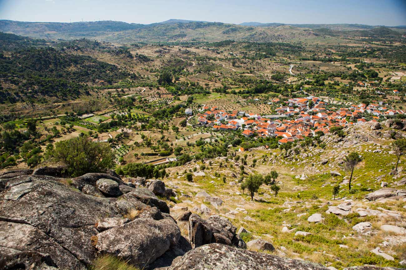 Full color slideshow photo showing a stunning view looking down on the village of Bendada, Portugal from a mountain side vista. Blue skies, green trees, pastures and mountain sides, and the terra-cotta roofs and white buildings of Bendada can be seen.