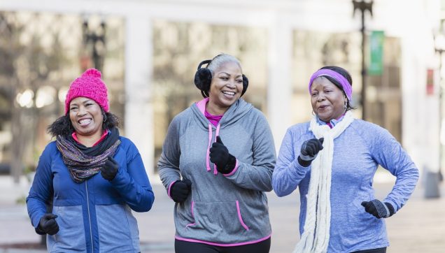 Three African-American women exercising together in the city, jogging or power walking, laughing and conversing. A building is out of focus in the background. The one on the left is in her 60s and her friends are mature women in their 40s and 50s. It is autumn or winter as they are wearing warm workout clothing, gloves, hats and ear muffs.
