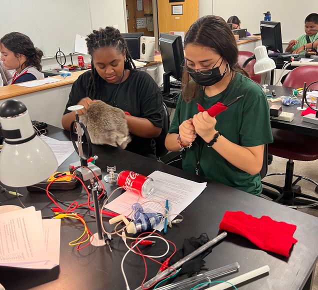 Two students sit at a lab bench and handle wires and a soda can.