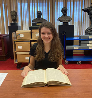 Catherine Devlin sitting at a table in front of an open book with busts in the background