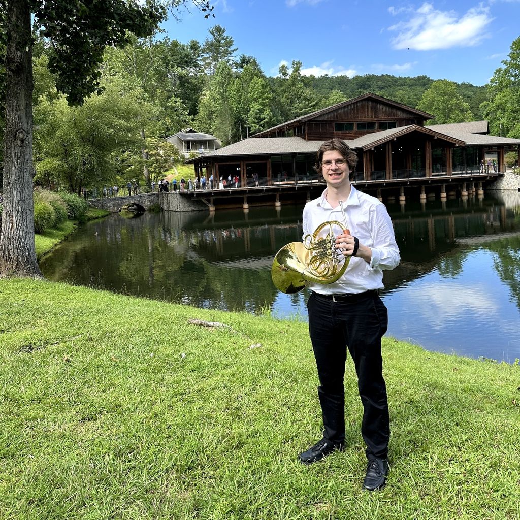 Chris Relyea, a BM Horn Performance and BA Computer Science dual degree student, is pictured holding a horn instrument and wearing a white long sleeve button down shirt with black pants. He is standing on grass and in the foreground, there is a lake and cabin-like venue, giving calming nature vibes.