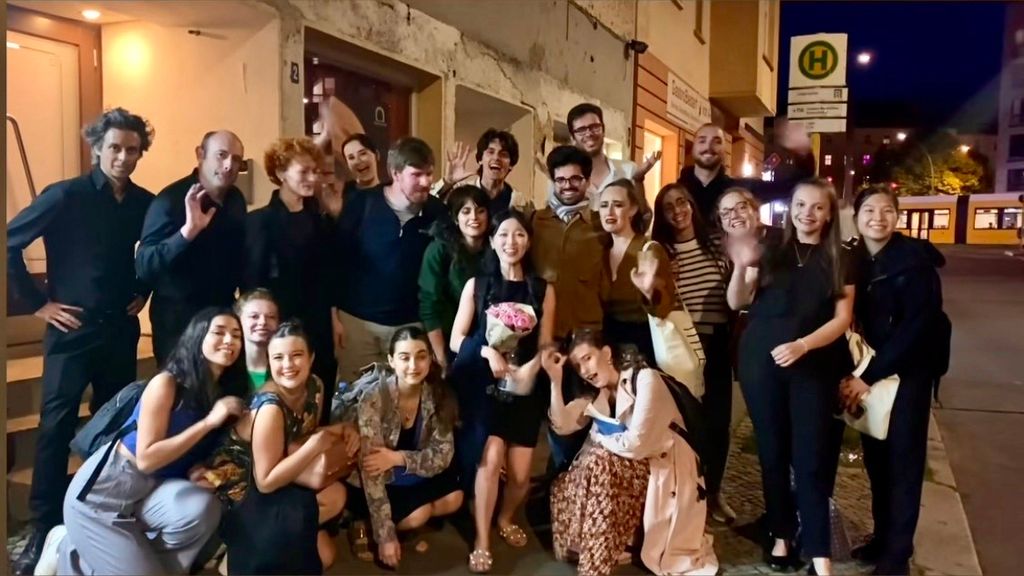 Group photo of a cheerful crowd gathered outside at night, posing and waving enthusiastically. A young woman in the center holds a bouquet of pink flowers, surrounded by smiling friends in casual and semi-formal attire. The setting appears to be a city street near a tram stop, with illuminated buildings in the background.